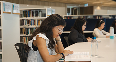 Student seated at desk in Library with a book and water bottle on the table in front of them