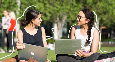 Two students with laptops sitting on grass chatting and smiling