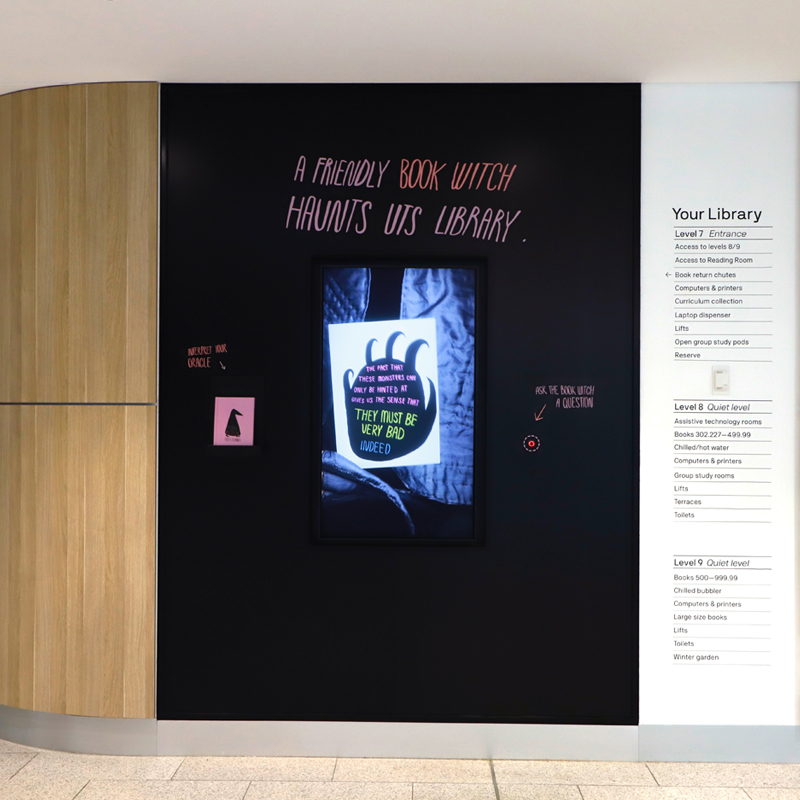 A screen sits centred on a black wall displaying a black clothed witch showing a tarot card. Above the screen 'A friendly BOOK WITCH haunts UTS Library' is written in pink and purple text