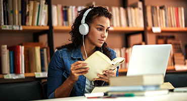 Lady at desk with earphones on looking at laptop and holding book in front of a book shelf