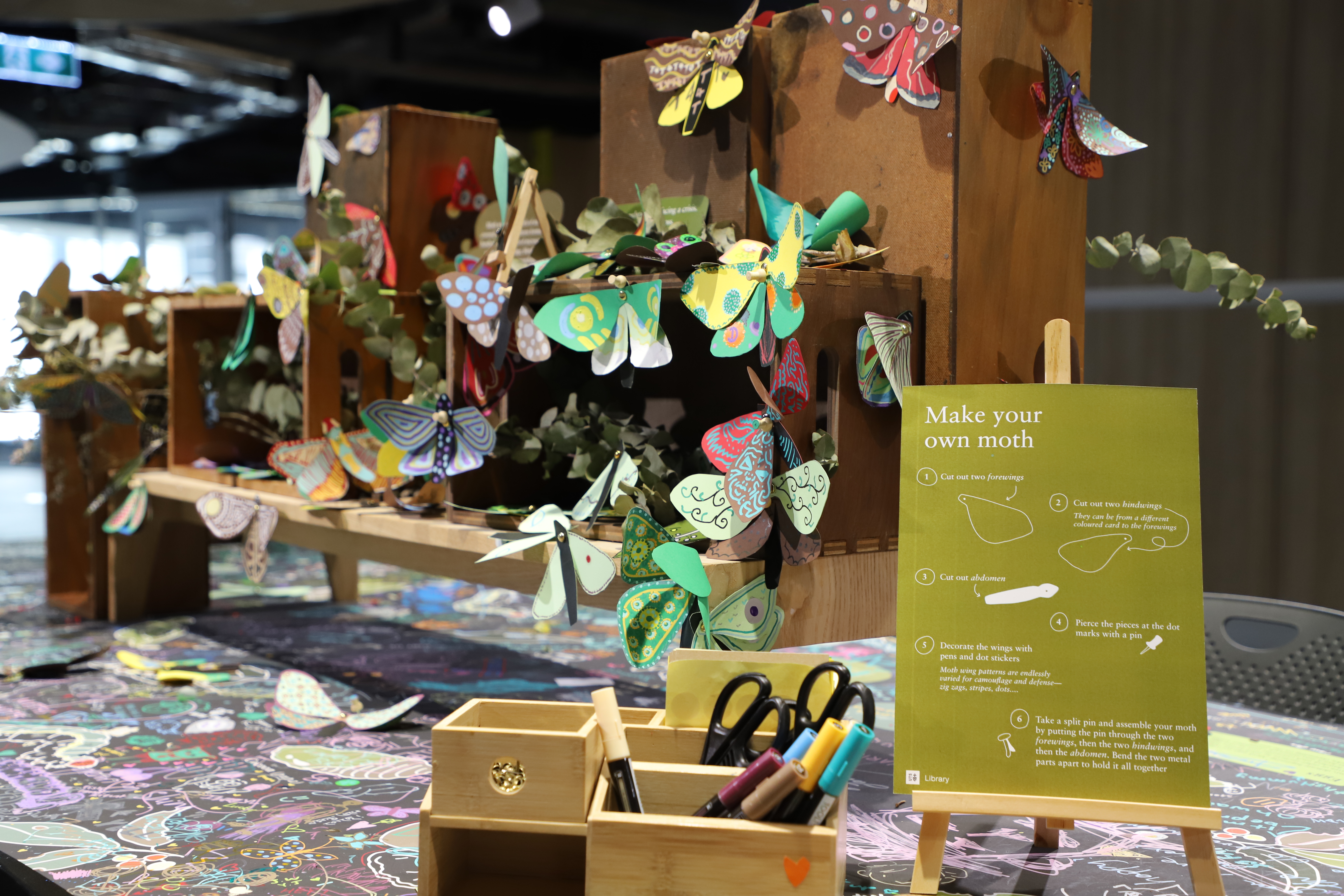 Coloured in paper moths are placed on wooden shelving. In front sits a sign promoting the creative activity