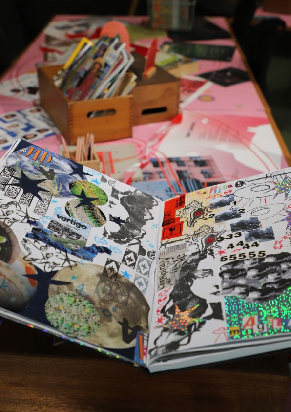 A large scrapbook filled with collages sits on top of a pink table, surrounded by craft supplies