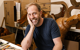 Simon Greiner in his studio, smiling and surrounded by cardboard and art‑making materials.