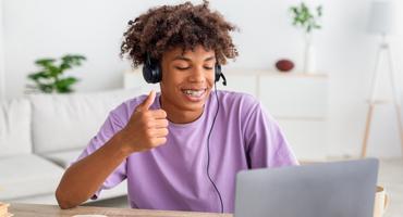 Boy in purple shirt with braces smiles and gives his laptop a thumbs up