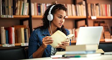 Lady at desk with earphones on looking at laptop and holding book in front of a book shelf