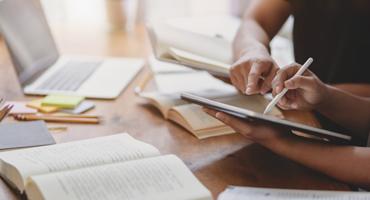 Students pointing at tablet, surrounded by books and a laptop