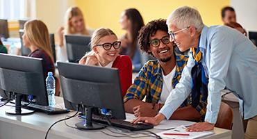 Students seated at a table with computers, trainer leaning over and pointing to something on the monitor, all smiling.