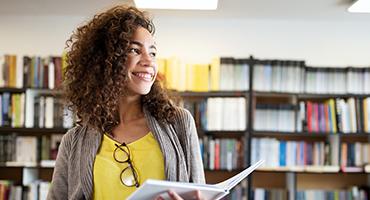 Woman in yellow top with an open book in her hand smiling and looking off into distance