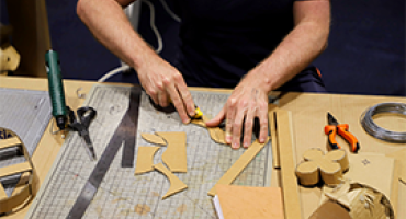 Hands cutting cardboard with workshop materials such as scissors, glue and cardboard scattered around them