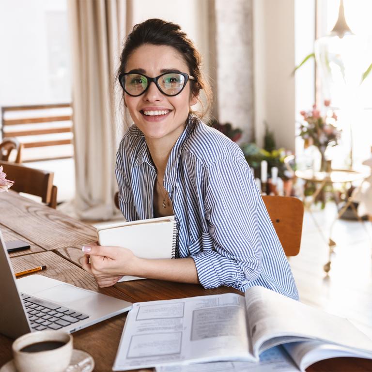Smiling woman seated at table in front of laptop, holding book