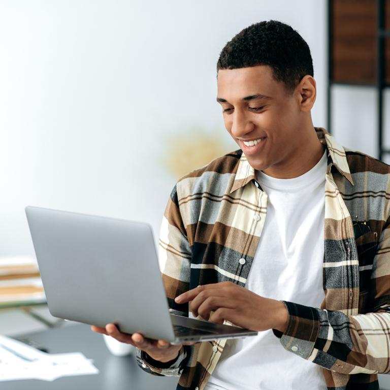 A boy in a green and white checkered shirt holds a silver laptop and looks at it smiling