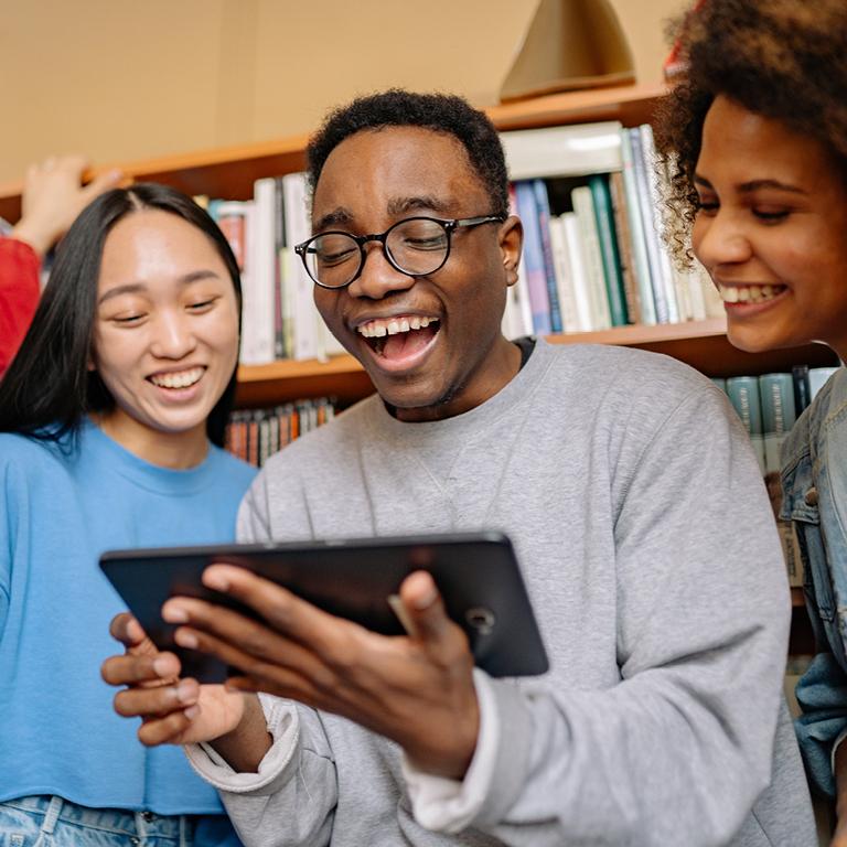 Three students gathered around a handheld tablet device and smiling gleefully