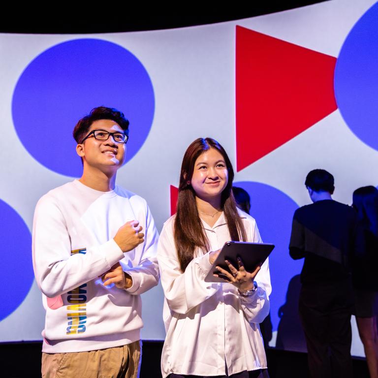 Two students smiling in the data arena, with a geometric pattern on the screen behind them