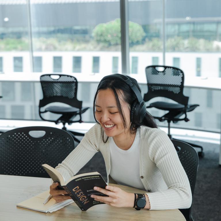 A female student in a white cardigan laughs as they read from a book