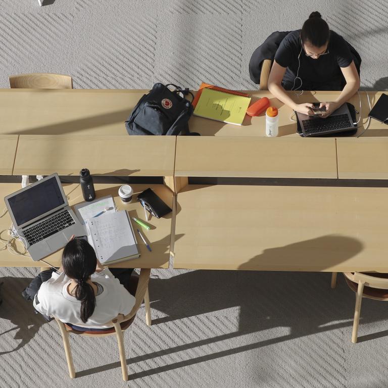 Overhead photo of students studying on wooden tables, laptops, books and pens scattered around them