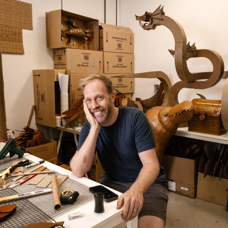 Blonde-haired artist smiles with face propped in one hand. Surrounding them are bits of cardboard and a cardboard dragon behind them.