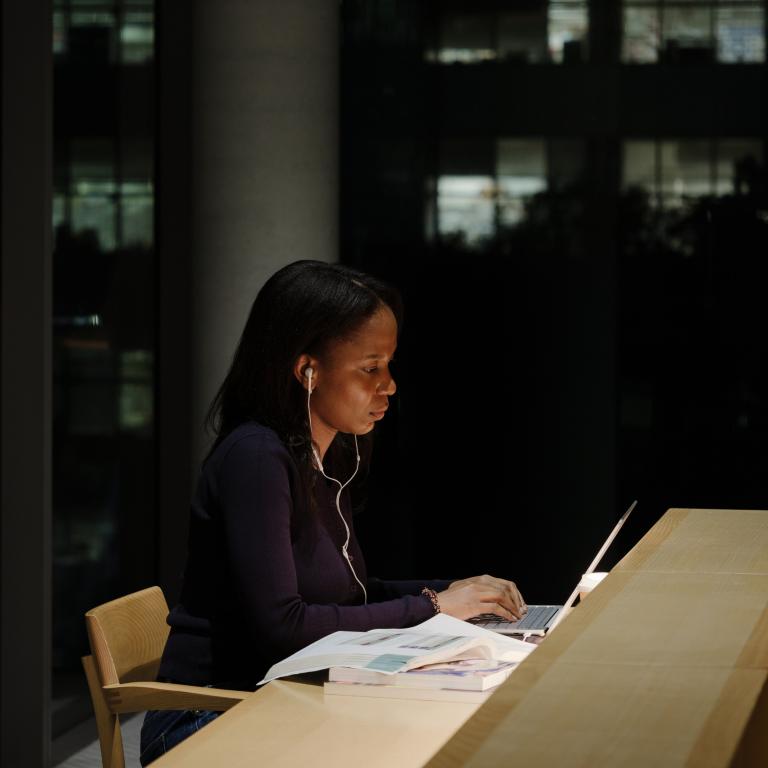 Women sits in at a wooden desk, working on a laptop. Behind her, are full glass windows showing the night sky and buildings behind her