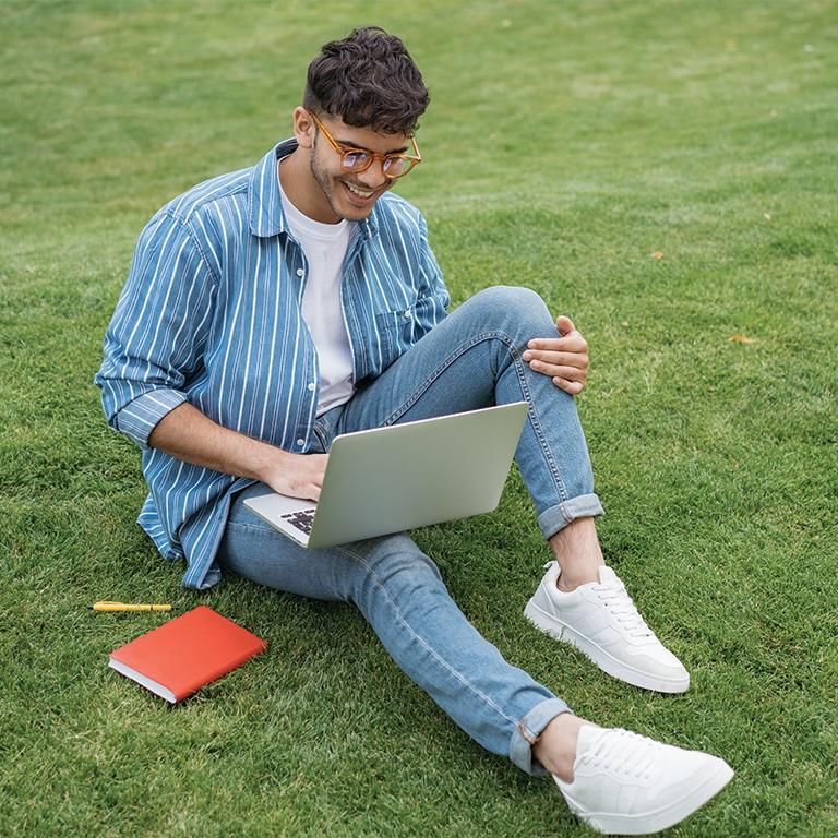 Smiling student sitting on grass looking at his laptop