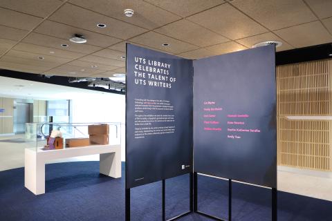 A photograph of two large mottled purple posters suspended on a black right-angle frame. These posters describe the exhibition and acknowledge key contributors