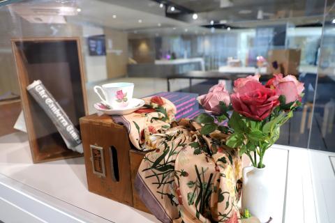 A photograph of inside one of the display cases in the exhibition. A tea cup sits poised on top of an old library catalogue card drawer which has been flipped on its side. A pink floral scarf cascades down towards a white vase with fake pink flowers