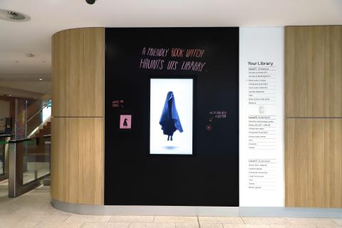 A black glass wall sits centred on a larger wall of wood. In the centre of the black wall, is a screen with a book witch floating inside. Above the screen reads 'A friendly BOOK WITCH haunts UTS Library'