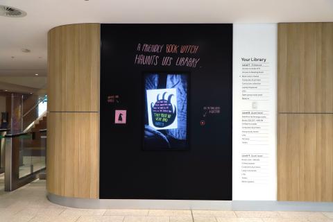 A black glass wall sits centred on a larger wall of wood. In the centre of the black wall, is a screen displaying a close-up of the BOOK WITCH's hands covered in claw-like fabric. She displays a tarot card. Above the screen reads 'A friendly BOOK WITCH haunts UTS Library'. 