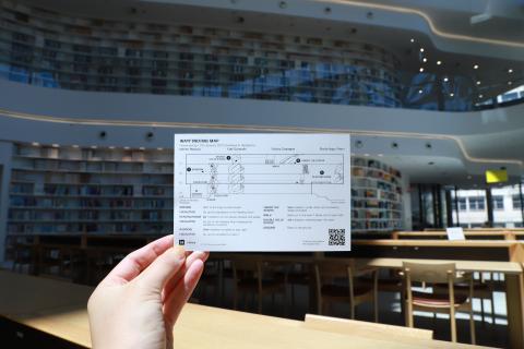 In the middle of a two-storeyed library with floor-to-ceiling bookshelves, a hand holds onto a postcard. The postcard has a map of the building, and instructs viewers on the key locations they can find installations of text around the building