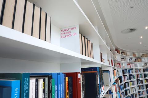 Up high on white bookshelves, pink text is stuck to a shelf end, stating 'Sometimes spaces silence us'
