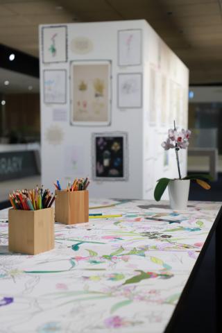 A table with boxes of pencils and an orchid in the foreground, with the exhibition wall with drawings hung up in the background. The table has a large colouring-in sheet on it that covers the whole surface.
