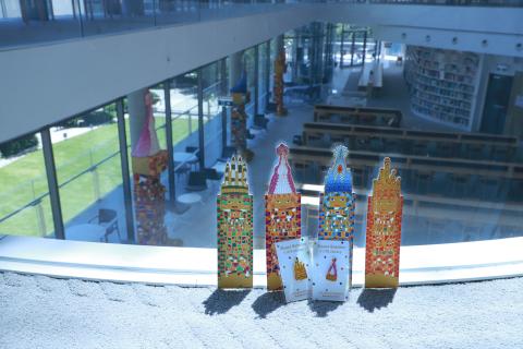 4 colourful bookmarks and 2 enamel pins sit placed against a glass bannister, looking down at their sculptural likenesses below