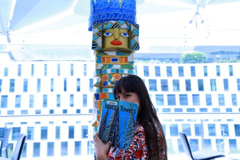 Creative in Residence Raquel Caballero holding a blue book to their face against a backdrop of their giant totem sculpture