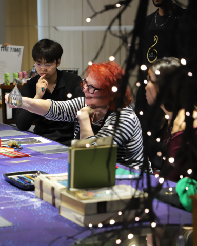 Woman leans on a table with one hand and holds up a collaged moth showing the others sitting at the table